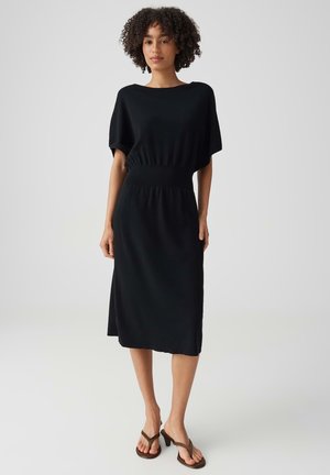 Donna con capelli ricci che indossa un vestito nero al ginocchio e sandali marroni con tacco, in piedi davanti a uno sfondo chiaro e uniforme.