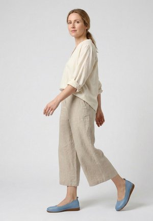 Kvinde gående i profil iført en løs cremefarvet bluse, beige bukser med brede ben og lyseblå flade sko mod en ensfarvet lys baggrund.