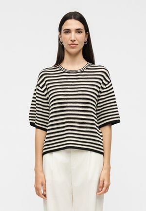 Γυναίκα με μακριά σκούρα μαλλιά, που φοράει μαύρο και άσπρο οριζόντια ριγέ πλεκτό κοντομάνικο τοπ και λευκό παντελόνι ψηλόμεσο, όρθια μπροστά σε μονόχρωμο φόντο.