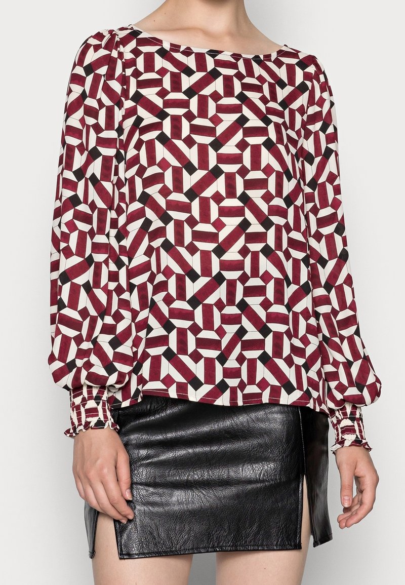 Femme portant un chemisier à manches longues avec un motif géométrique en bordeaux, noir et blanc, associé à une mini-jupe en cuir noir avec des fentes latérales.