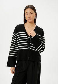Schwarz-weiß gestreifter Cardigan mit langen Ärmeln, V-Ausschnitt und fünf runden Knöpfen vorne. Weicher Stoff, kurzes Design.