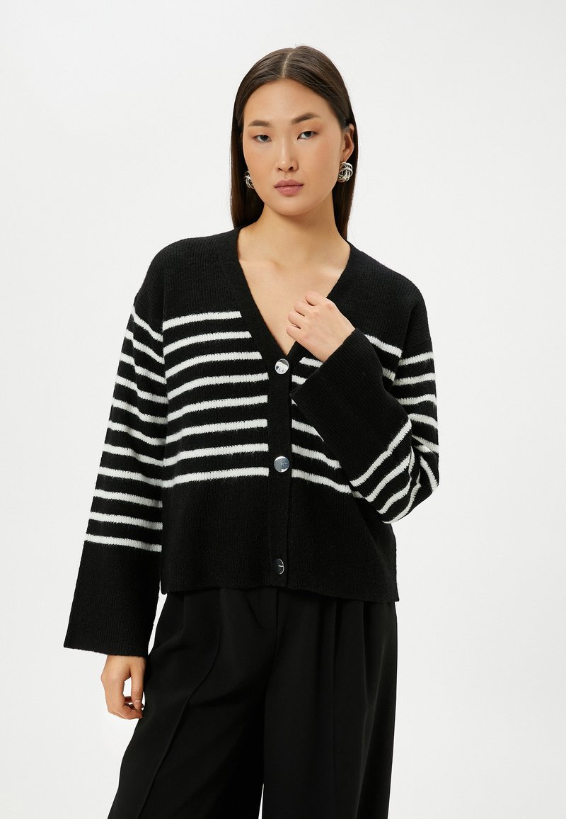 Schwarz-weiß gestreifter Cardigan mit langen Ärmeln, V-Ausschnitt und fünf runden Knöpfen vorne. Weicher Stoff, kurzes Design.