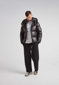 Giubbotto piumino nero con cappuccio, materiale lucido, design trapuntato, indossato sopra una maglietta grigia, abbinato a pantaloni della tuta neri e sneakers.