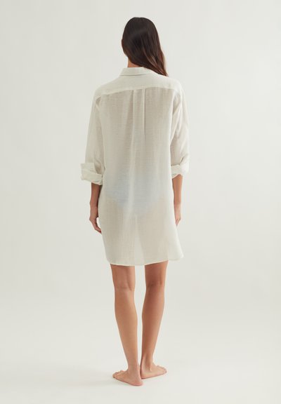 Γυναίκα όρθια, ξυπόλητη, γυρνισμένη με την πλάτη, φοράει ένα διαφανές, λευκό φόρεμα με μακριά μανίκια που φτάνει μέχρι τα γόνατα και τα μανίκια είναι γυρισμένα.