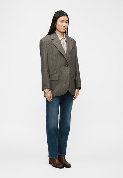 Νεαρή γυναίκα με μακριά σκούρα μαλλιά, που φοράει υπερμεγέθη καφέ καρό σακάκι, μπεζ πουκάμισο, μπλε τζιν και καφέ μποτάκια μέχρι τον αστράγαλο, στέκεται μπροστά σε λευκό φόντο.