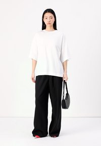 Witte oversized t-shirt met korte mouwen, gecombineerd met brede zwarte broek, met rode accenten bij de manchetten, en een kleine zwarte tas met perforaties.