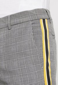 Šedé kárované kalhoty s texturovaným vzorem, boční pruhy ve žluté a černé barvě, s upraveným střihem a detaily kapes.