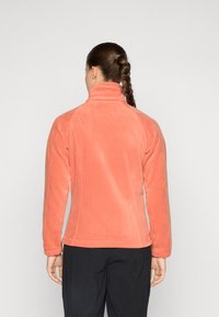 Veste en polaire de couleur pêche avec un col haut, une coupe ajustée et des manches longues. Texture lisse et détails de couture visibles.