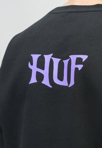 Vista traseira de uma pessoa vestindo uma peça de vestuário preta com a palavra "HUF" impressa em grandes letras góticas roxas na parte superior das costas.