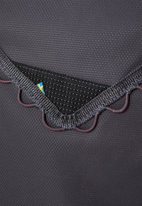 Tissu noir texturé avec une couche intérieure à motifs, présentant des coutures décoratives grises et rouges le long des bords et une petite étiquette bleue.
