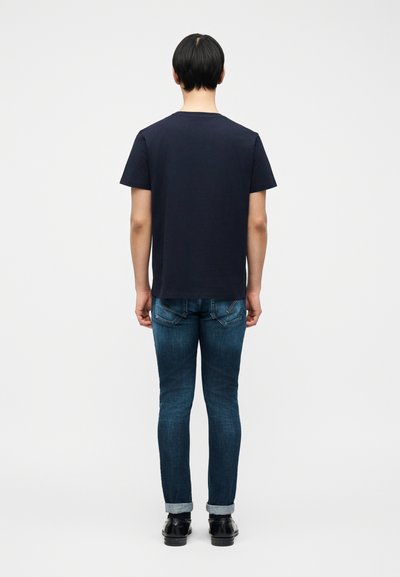 Άνδρας που στέκεται γυρισμένος με την πλάτη, φοράει απλό μαύρο μπλουζάκι, σκούρα μπλε τζιν με μανσέτες και μαύρα επίσημα παπούτσια σε λευκό φόντο.