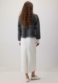 Ženska z dolgimi curly lasmi, oblečena v temno sivo pulover, belo dolgo krilo z zarezo na hrbtu in bele čevlje, stoji ob navadni steni.