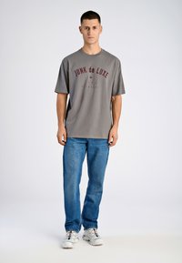 Camiseta gris con el logo "JUNK de LUXE" en marrón, corte relajado, combinada con jeans de mezclilla azul claro y zapatillas blancas. Fondo sencillo.
