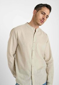 Man in een beige overhemd met mandarin kraag, staand tegen een witte achtergrond, kijkend naar beneden en iets opzij.