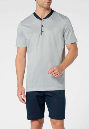 Uomo che indossa una maglietta henley a maniche corte di colore grigio chiaro con colletto e bottoni neri, abbinata a pantaloncini blu scuro, in piedi contro uno sfondo bianco.