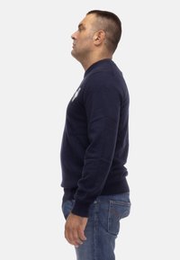 Maglione blu navy con collo rotondo e maniche lunghe, caratterizzato da un sottile dettaglio del logo sul lato sinistro. Tessuto morbido con un orlo aderente.