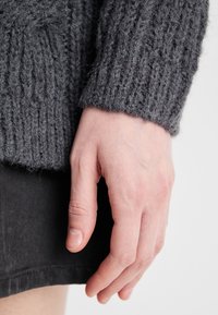 Grauer gestrickter Pullover mit einem strukturierten Rippmuster am Bündchen, mit einer Nahaufnahme einer Hand vor einem dunklen Stoffhintergrund.