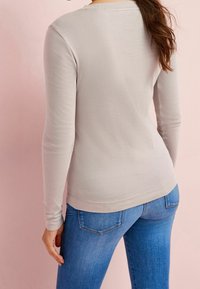 Camicia a maniche lunghe beige chiaro con scollatura rotonda, design semplice e tessuto elasticizzato, abbinata a jeans blu in denim mostrati sul retro.