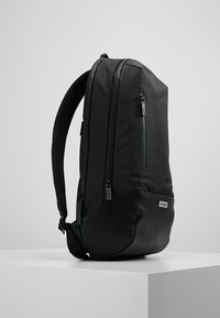Zaino nero elegante con molteplici scomparti con cerniera, pannello posteriore imbottito e base curvata, esposto su un piedistallo bianco in un contesto neutro.