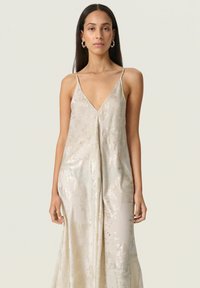 Woman with long dark hair wears sleeveless, light beige satin dress with subtle metallic pattern and thin shoulder straps, standing against plain background.