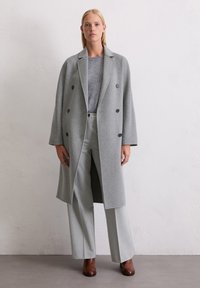 Grauer Woll-Doppelreiher mit Reverskragen, lockerer Passform und Knopf-Details, getragen über einem grauen Pullover und hellgrauen Hosen. Braune Stiefeletten.