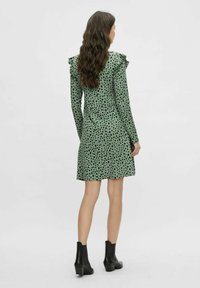 Zelené šaty s černými puntíky, dlouhé rukávy, na ramenou volány, do délky ke kolenům, nošeny s černými kotníkovými botami. Hladká textura látky.