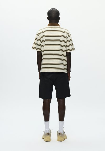 Polo rayé beige et marron, short noir, chaussettes blanches et baskets beiges. Modèle vu de dos sur un fond uni.
