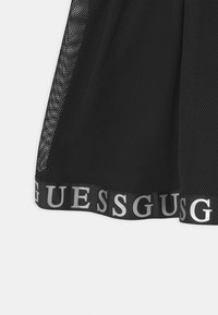Černá síťovaná sukně s širokým elastickým pasem a opakujícím se logem "GUESS" v stříbrné barvě. Lehký, prodyšný materiál s texturovaným povrchem.