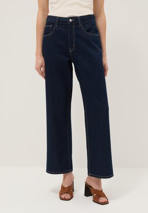 Mujer con vaqueros de pierna ancha de talle alto en azul oscuro con costuras claras, camiseta blanca y sandalias de tacón abiertas marrones sobre un fondo neutro.