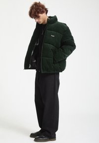 Green corduroy puffer jacket with high collar, zip closure, and side pockets. Worn over a black shirt and paired with black trousers and shoes.