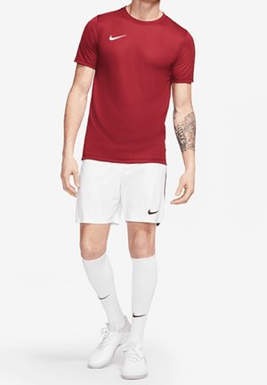 Mężczyzna w czerwonej sportowej koszulce Nike, białych szortach Nike, białych skarpetach do kolan Nike i białych butach sportowych, stoi z jedną nogą lekko wysuniętą do przodu.