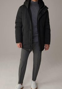 Schwarze Steppjacke mit Kapuze, ausgestattet mit einem Reißverschluss und Knopfverschluss. Getragen über einem grauen Rollkragenpullover und kombiniert mit grauen Hosen und weißen Sneakers.