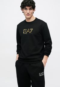 Svart genser med ribbestrikkede mansjetter og kant, med en gullfarget EA7 Emporio Armani-logo på brystet, kombinert med matchende svarte joggebukser.