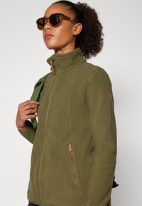 Olive rohelise fleece-jakk kõrge kraega, esiküljel pikitad ja pruun nahast detailid; mudel kannab päikeseprille ja hoiab rohelist rihma.