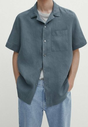 Man wearing a short-sleeve, button-up dark gray shirt over a white t-shirt, paired with light blue jeans, hands in pockets.