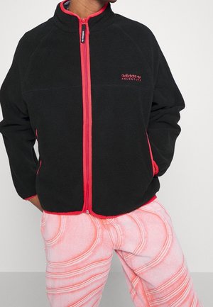 Osoba nosząca czarną kurtkę polarową Adidas Adventure z czerwonym zamkiem i obszyciem, w połączeniu z jasnoróżowymi spodniami z czerwonymi wzorami w kształcie wirów.