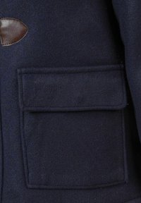 Mornarska jakna od vune s dodatkom, koja sadrži kožnu oznaku na ramenu i veliki prednji džep s poklopcem. Mekana tekstura i strukturirani dizajn.