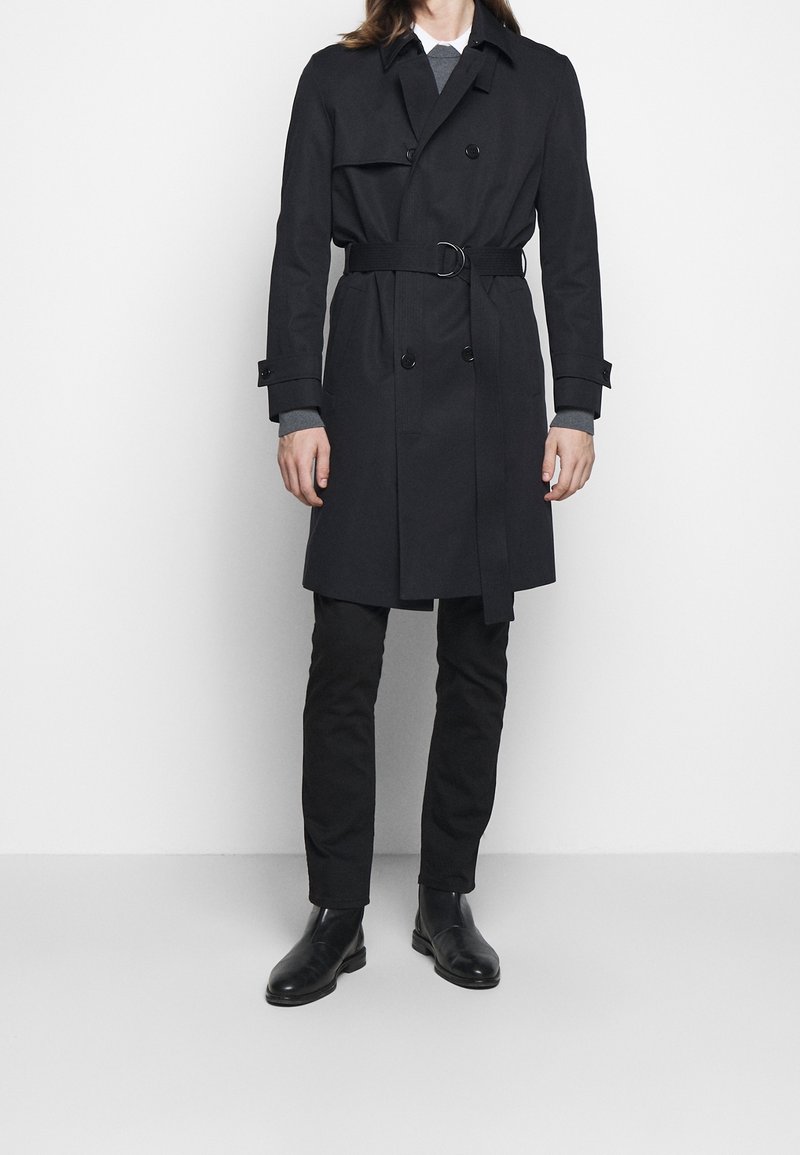 Personne portant un trench-coat noir ceinturé par-dessus un pull gris et une chemise à col, un pantalon noir et des bottines en cuir noir, debout.