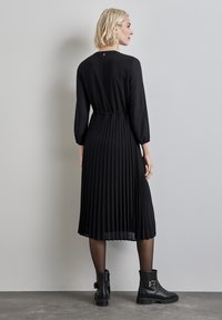 Schwarzes plissiertes Kleid mit langen Ärmeln, tailliert und aus strukturiertem Stoff. Kombiniert mit schwarzen Stiefeletten und transparenten Strumpfhosen.