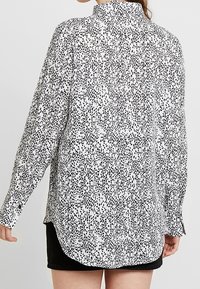 Långärmad skjorta med hög krage, tillverkad av ett lätt material, med ett svart och vitt prickigt mönster och välvd nederkant.