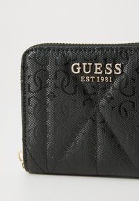 Portefeuille noir avec un design texturé et le logo "GUESS" en or. Il dispose d'une fermeture éclair et de détails en motif géométrique.