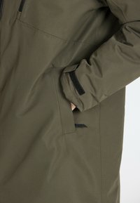 Donker olijfgroen outerwear met een gladde, gestructureerde afwerking, voorzien van een zijzak en zwarte klittenbandaccenten op de mouwen.