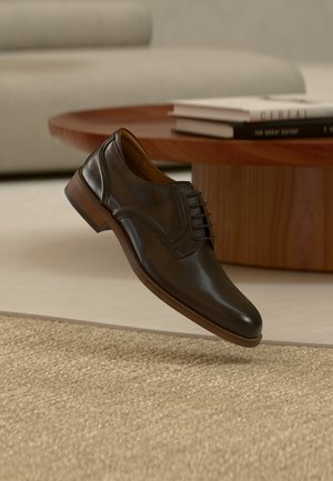 Dark brown leather dress shoe with laces floating above a beige woven rug, wooden coffee table, and light-colored sofa in the background.