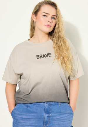 T-shirt w gradientzie od beżu do szarości z napisem "BRAVE" w kolorze czarnym; krótkie rękawy, luźny krój. W zestawie z jasnoniebieskimi dżinsami.