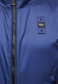 Fermeture éclair de veste bleue avec des dents noires, un écusson cousu portant l'inscription « Blauer U.S.A. » avec deux œillets métalliques sur la zone de la poitrine.