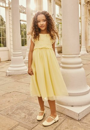 Gelbes Kleid mit Blumenakzenten und Rüschenärmeln, mehrlagigem Rock, kombiniert mit hellgelben Mary-Janeschuhen. Im Hintergrund weiße Säulen.