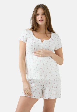 Fehér virágmintás rövid ujjú felsőt és hozzáillő rövidnadrágot viselő terhes nő, aki gyengéden tartja a hasát, egyszínű háttér előtt áll.