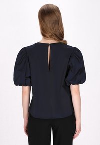 Marineblaue Bluse mit Puffärmeln und Schlüssel-Rücken, aus glattem Stoff gefertigt. Das Design verfügt über einen runden Ausschnitt und eine taillierte Silhouette.
