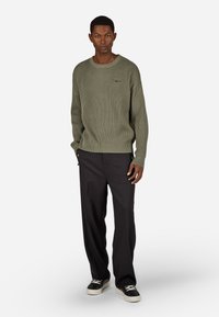 Oliwkowy sweter z prążkowanej tkaniny, z długimi rękawami i okrągłym dekoltem, w zestawieniu z czarnymi spodniami i czarnymi trampkami z białymi podeszwami.