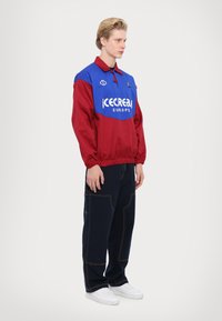 Giacca pullover rossa e blu con colletto e vita elastica, caratterizzata da un logo bianco "icecream EUROPE" e pantaloni blu navy con cuciture a contrasto.